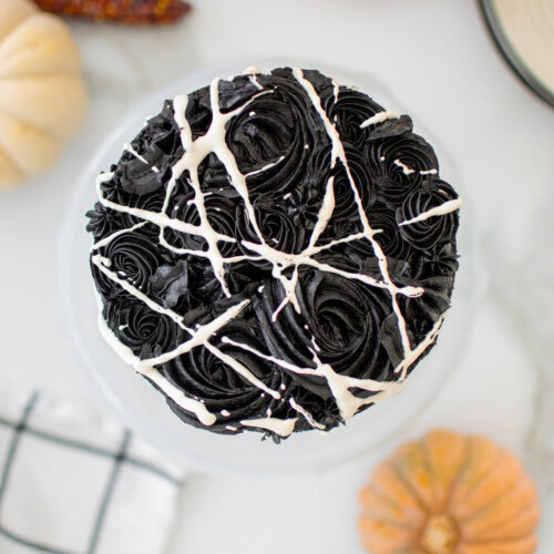 spooky rose cake for halloween on a cake stand on a white marble counter with pumpkins.