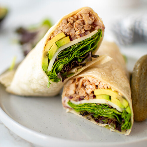 bbq turkey avocado wrap cut in half on a plate with pickles on a white marble countertop.