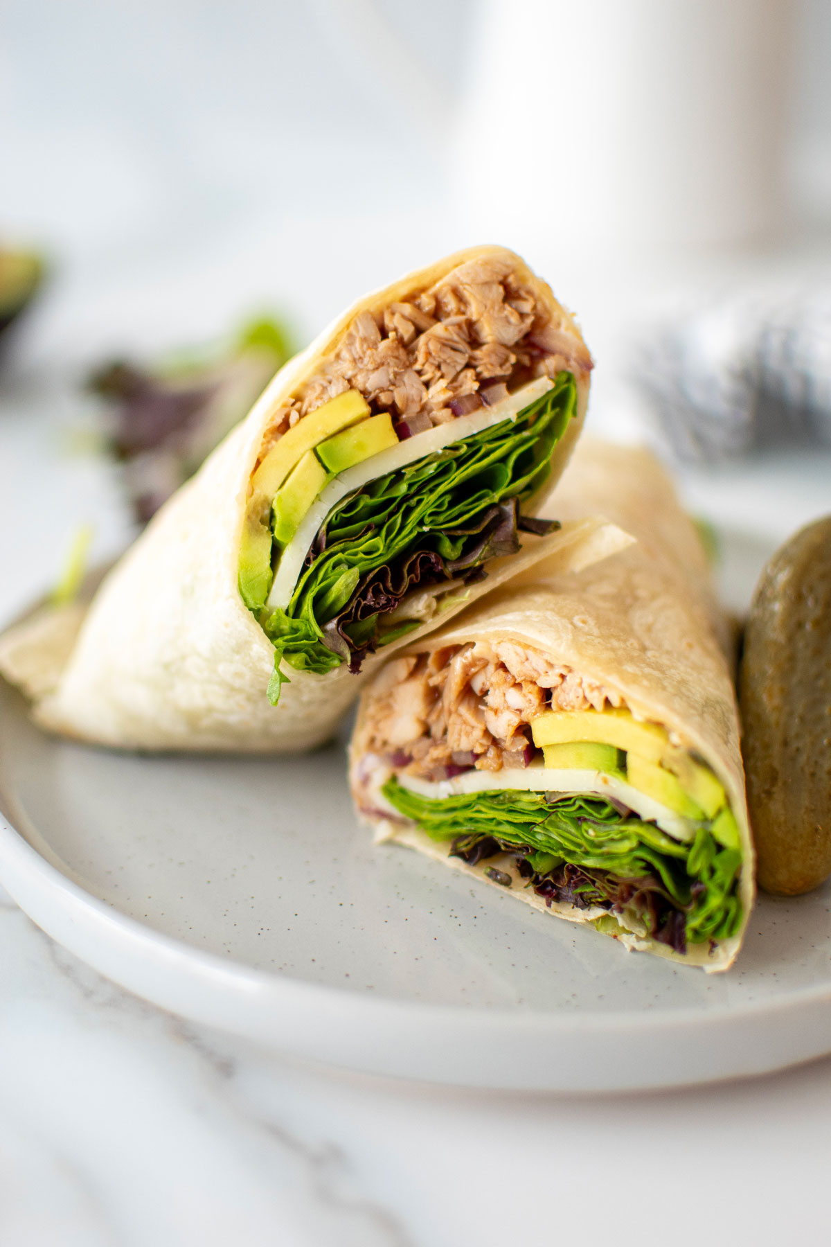 bbq turkey avocado wrap cut in half on a plate with pickles on a white marble countertop.
