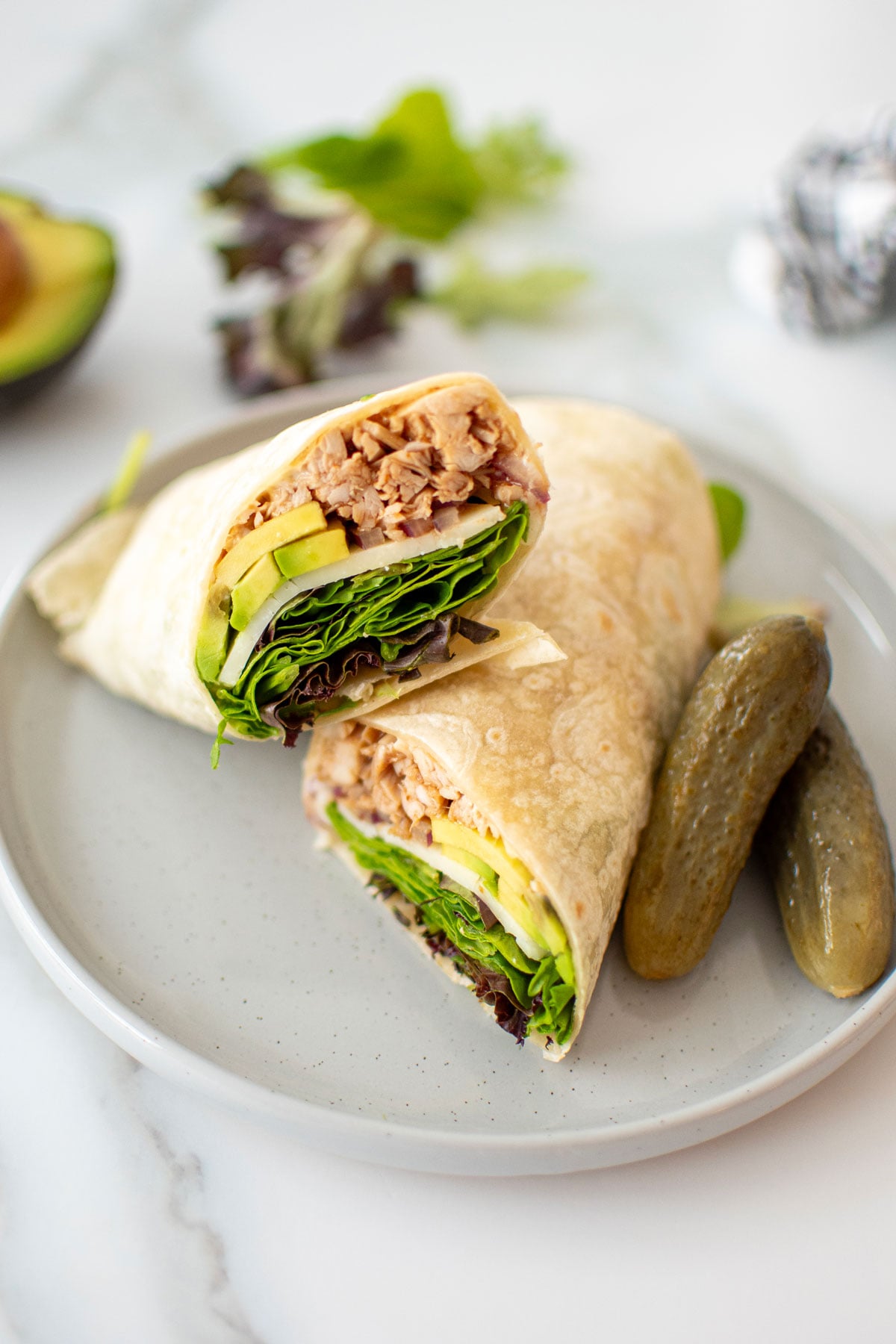 bbq turkey avocado wrap cut in half on a plate with pickles on a white marble countertop.