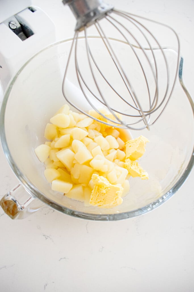 cooked potatoes, bone broth and butter in a glass mixing bowl with a whisk on a marble counter.