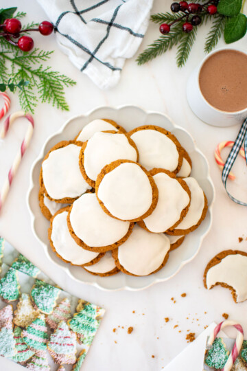 plate of gluten free frosted gingerbread cookies on a white marble counter with a mug of hot cocoa, holiday cards and candy canes.