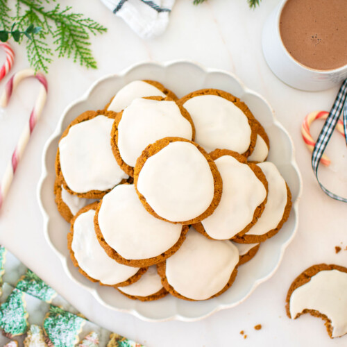 plate of gluten free frosted gingerbread cookies on a white marble counter with a mug of hot cocoa, holiday cards and candy canes.
