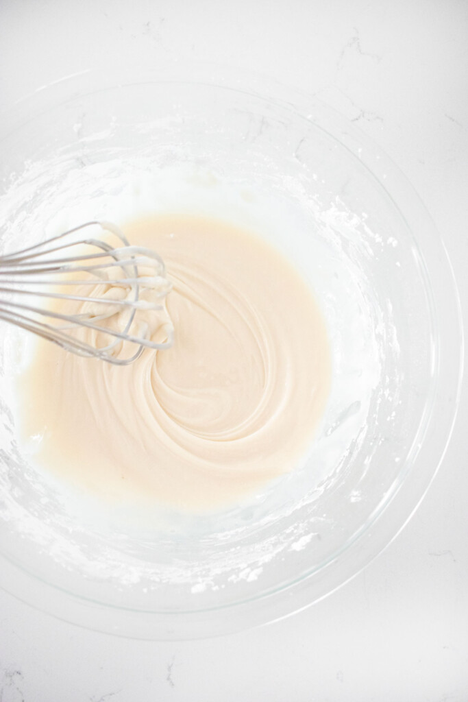 icing in a glass mixing bowl with a whisk on a white marble counter.