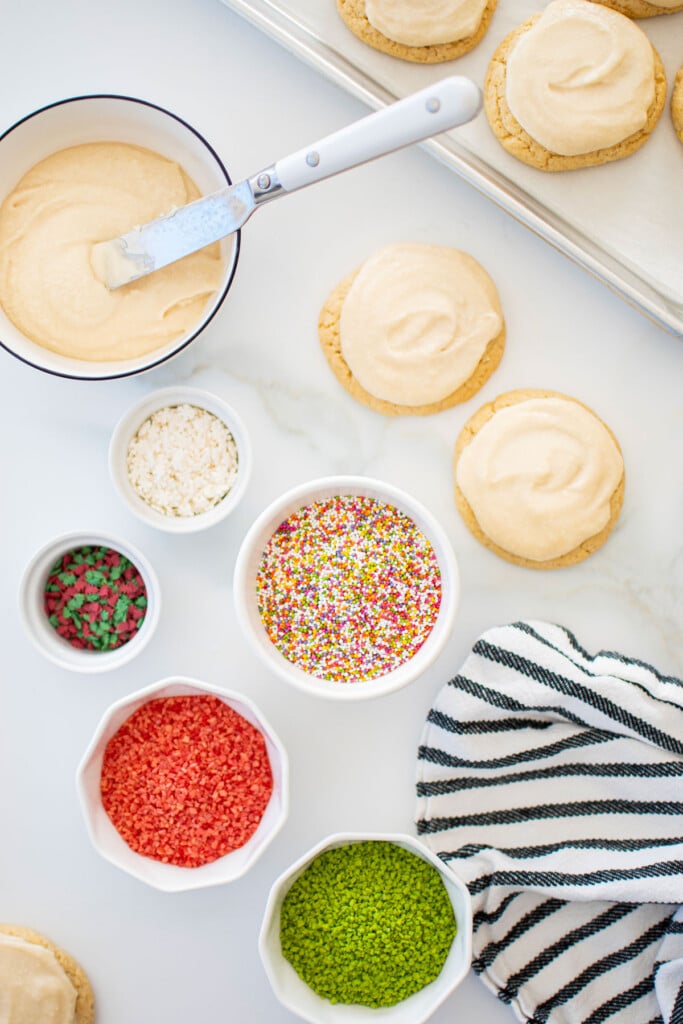 sugar cookies getting decorated with icing and sprinkles on a white marble counter.