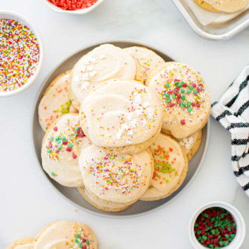 plate of holiday sugar cookies with icing and bowls of sprinkles on a white marble counter.