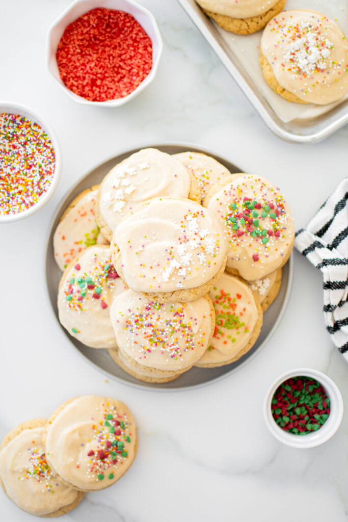 plate of holiday sugar cookies with icing and bowls of sprinkles on a white marble counter.