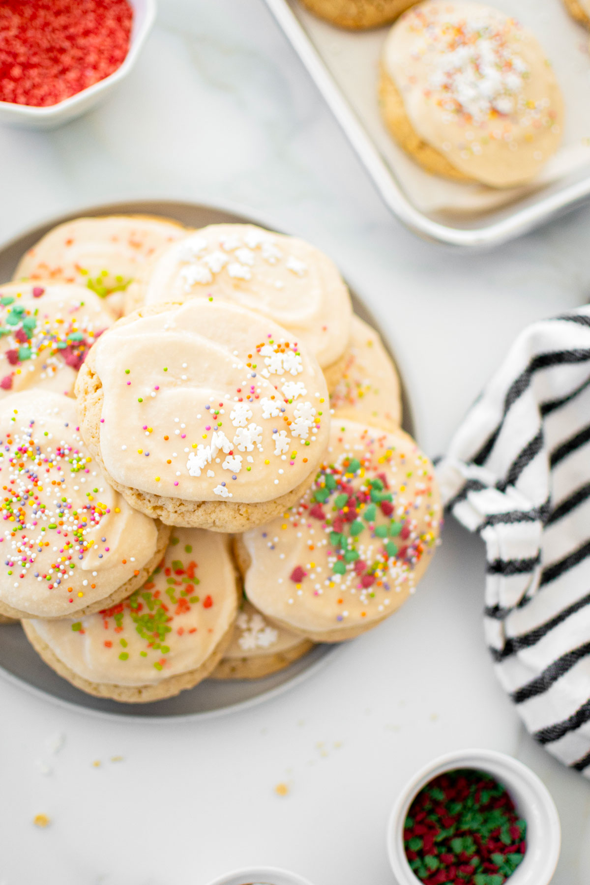 plate of holiday sugar cookies with icing and bowls of sprinkles on a white marble counter.