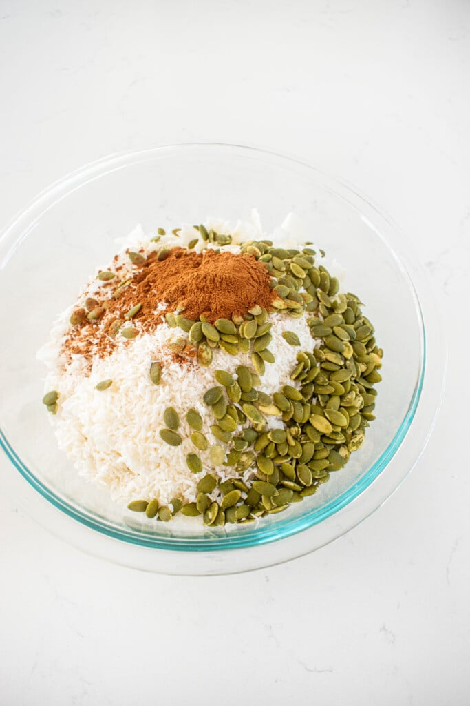 dry ingredients for maple cinnamon olive oil granola in a glass mixing bowl on a white marble counter.