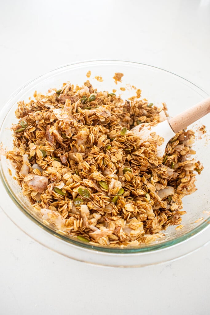 ingredients being mixed for maple cinnamon olive oil granola in a glass mixing bowl on a white marble counter.