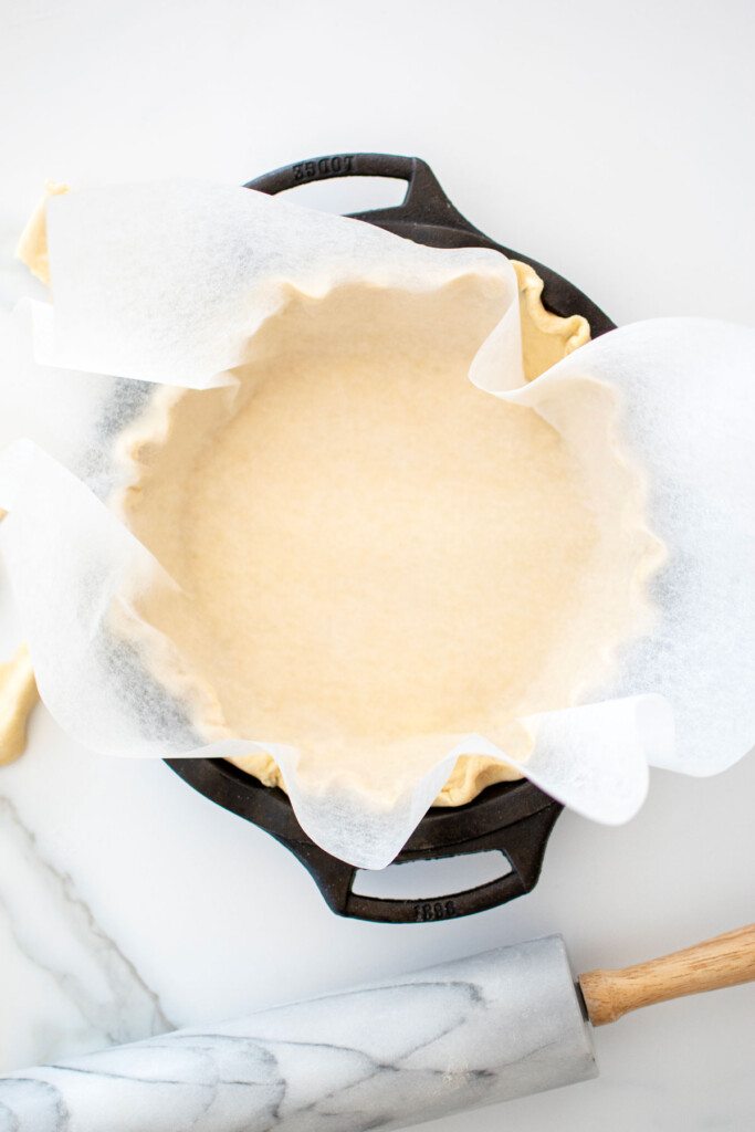 pie crust in a cast iron pie pan with parchment paper on a white marble counter.