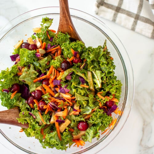 simple cranberry kale salad with homemade dressing in a glass bowl on a white marble countertop with wood salad spoons.