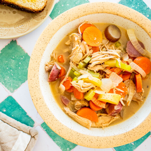 turkey carcass soup with bone broth in a bowl with a side plate of bread and butter on a green tile table.