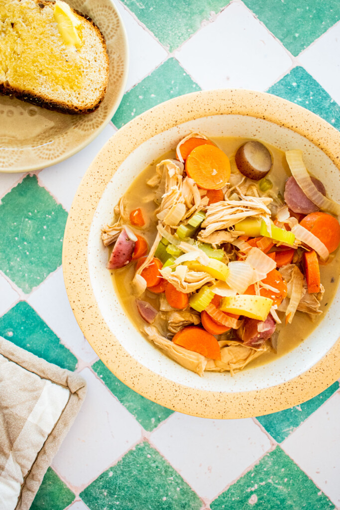 turkey carcass soup with bone broth in a bowl with a side plate of bread and butter on a green tile table.