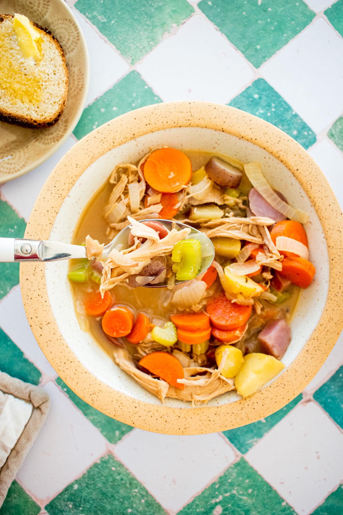 turkey carcass soup with bone broth in a bowl with a side plate of bread and butter on a green tile table.