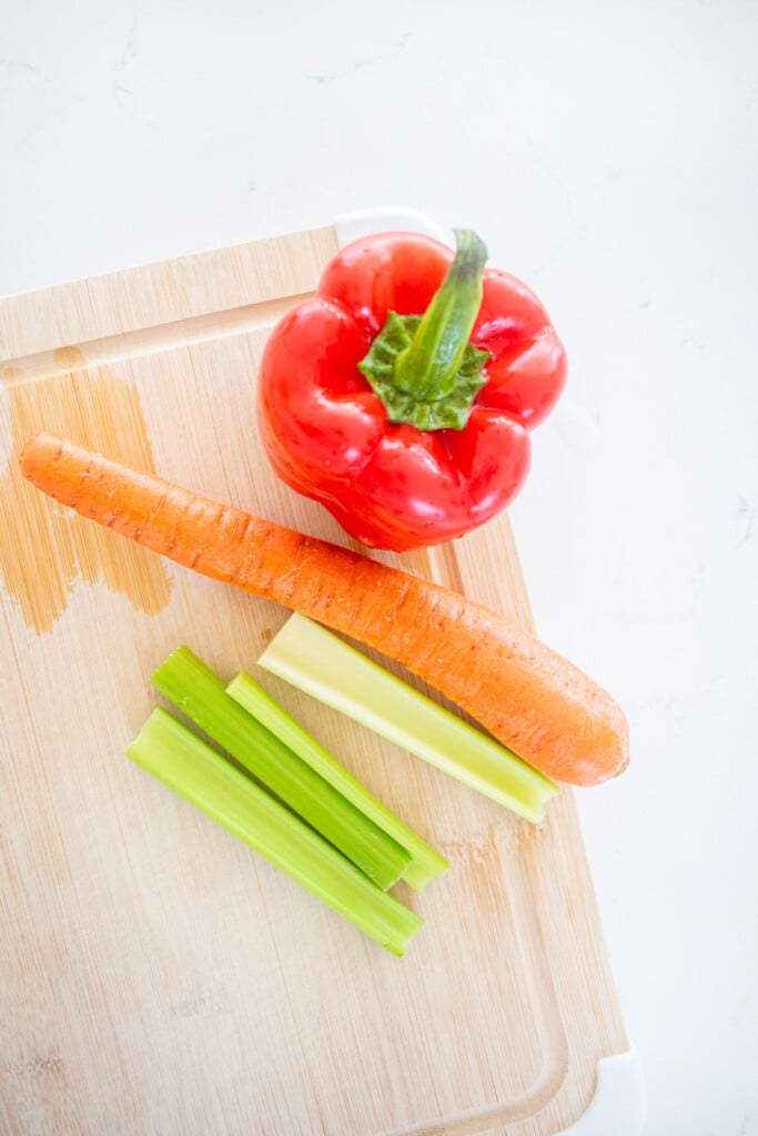 wood cutting board with red bell pepper, carrot and celery on a white marble countertop.