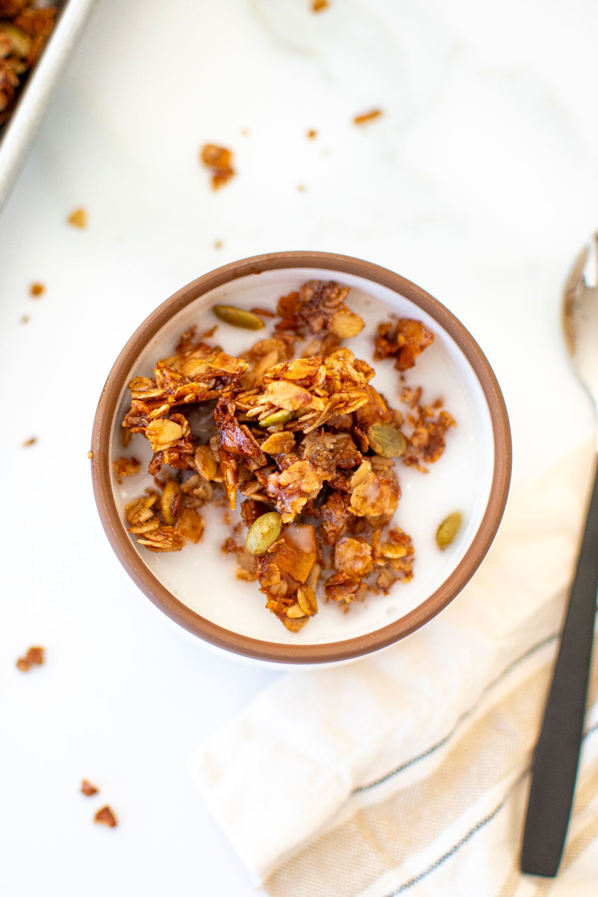 maple cinnamon olive oil granola in a bowl with milk on a white marble counter.