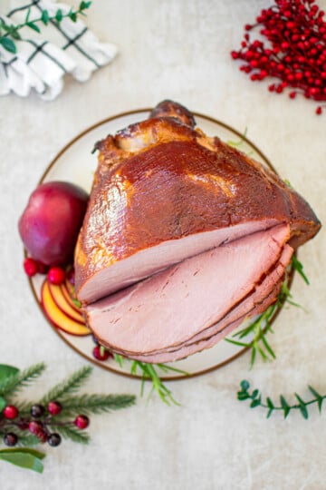 apple cider honey baked ham cut into slices on a plate garnished with apple, cranberries and fresh rosemary.