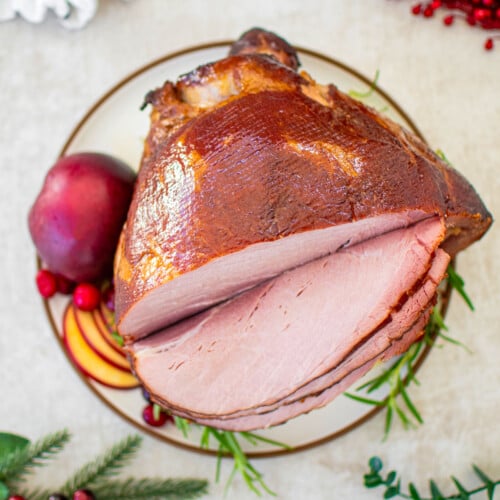 apple cider honey baked ham cut into slices on a plate garnished with apple, cranberries and fresh rosemary.