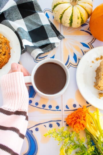 woman holding a mug of bone broth hot cocoa on a tile table with fresh flowers, cookies, a slice of cake on a plate and tiny pumpkins.