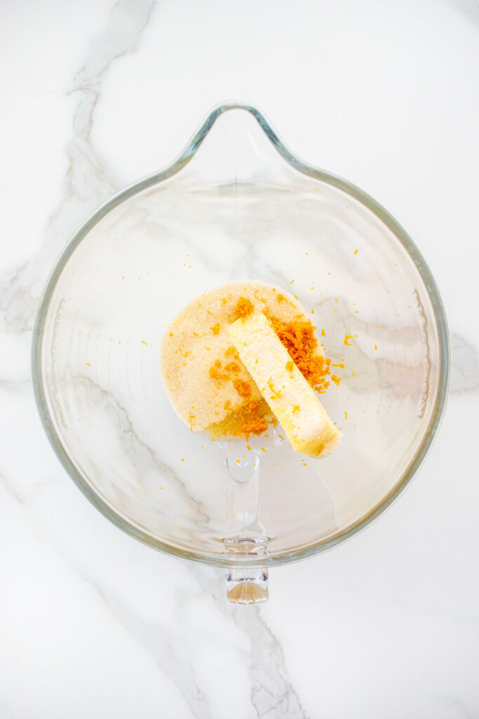 butter sugar and orange zest in a glass mixing bowl on a marble counter.