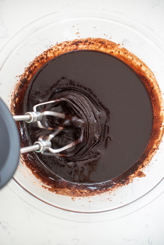 brownie batter being mixed with a hand mixer in a glass bowl on a marble counter.