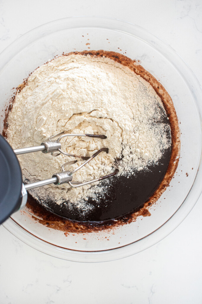 brownie batter being mixed with a hand mixer in a glass bowl on a marble counter.