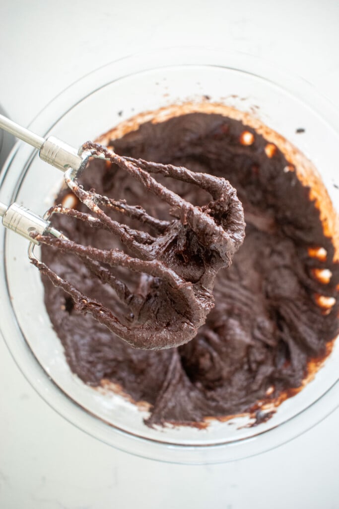 brownie batter being mixed with a hand mixer in a glass bowl on a marble counter.