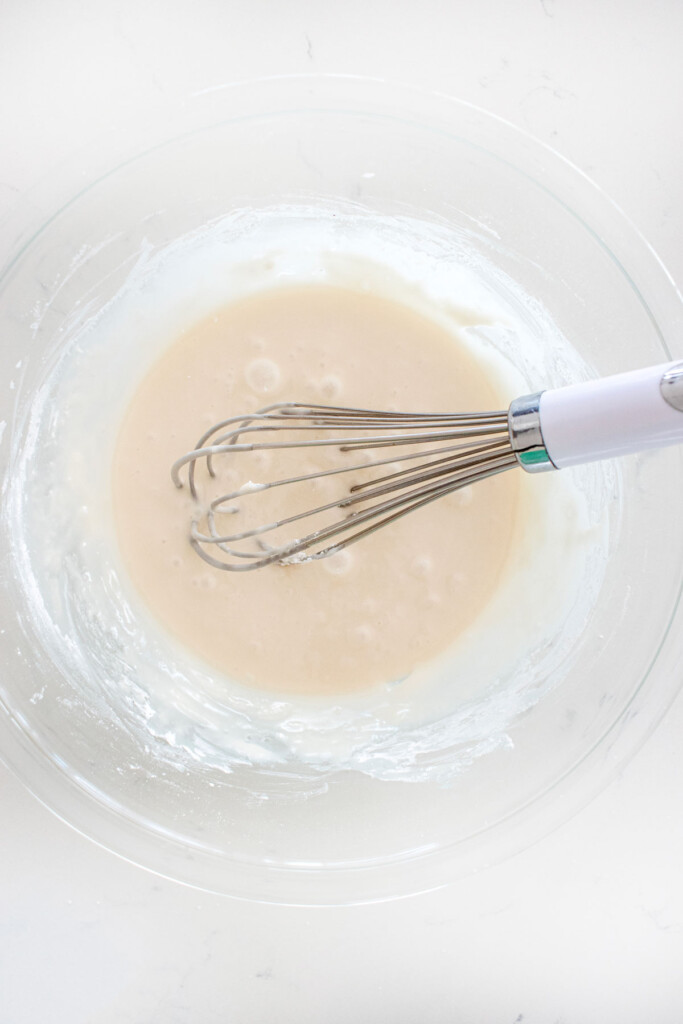 white glaze in a glass mixing bowl with a whisk on a marble counter.