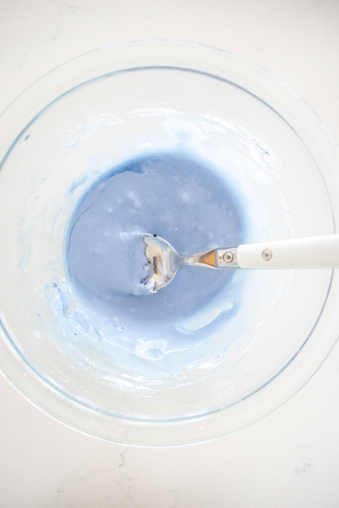 light blue decorating glaze in a glass mixing bowl on a white marble counter.