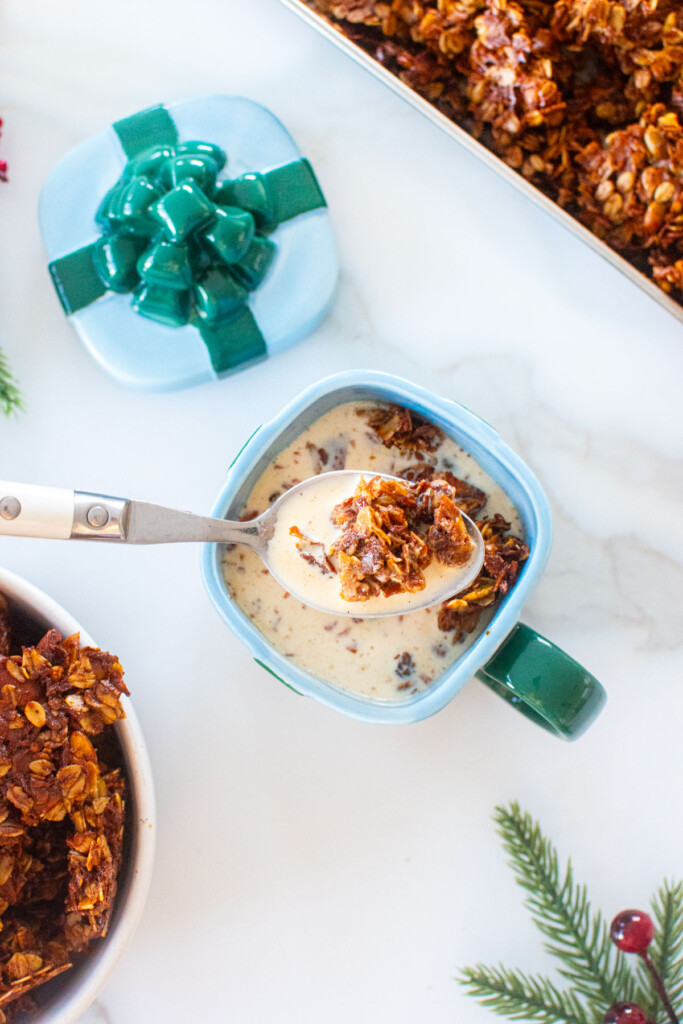 gingerbread granola in a present mug with milk and a spoon on a white marble counter.