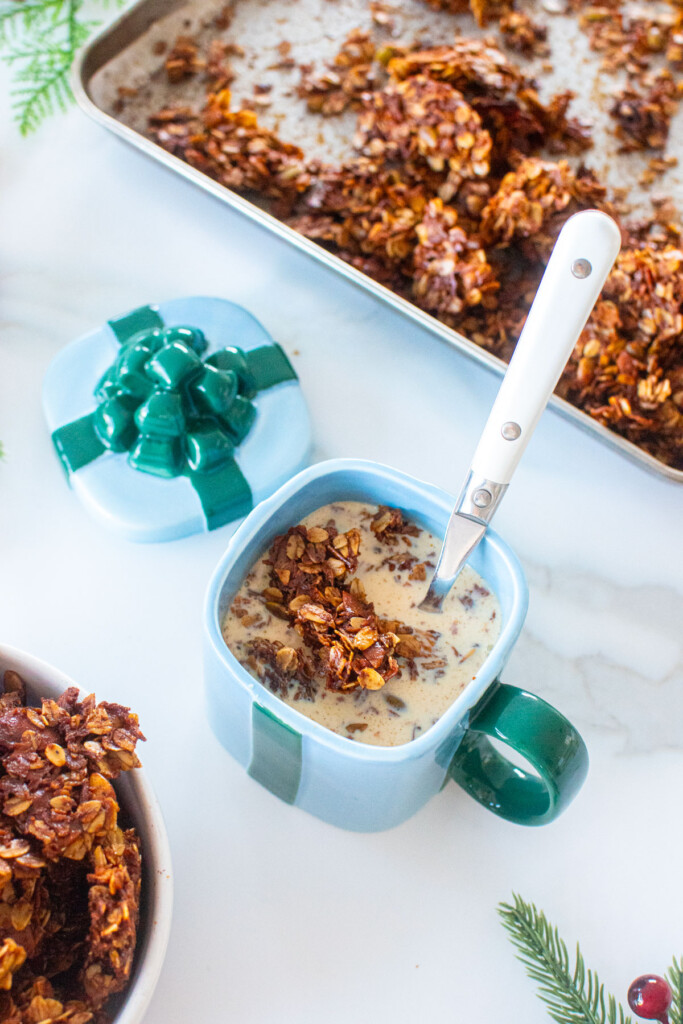 gingerbread granola in a present mug with milk and a spoon on a white marble counter.