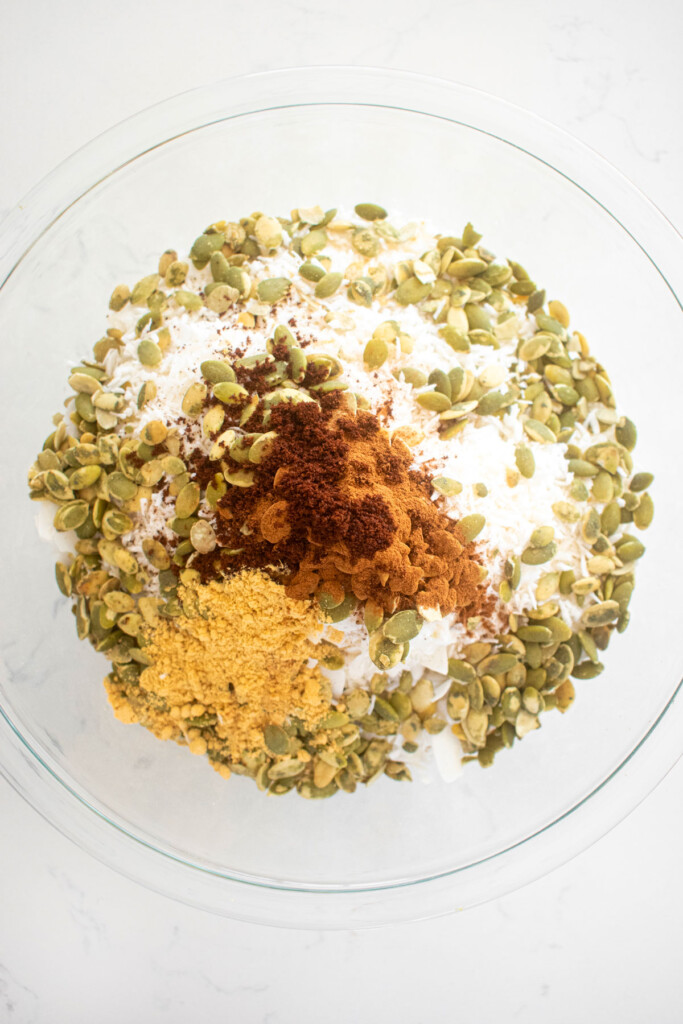 ingredients for healthy gingerbread granola in a glass mixing bowl on a white marble counter.