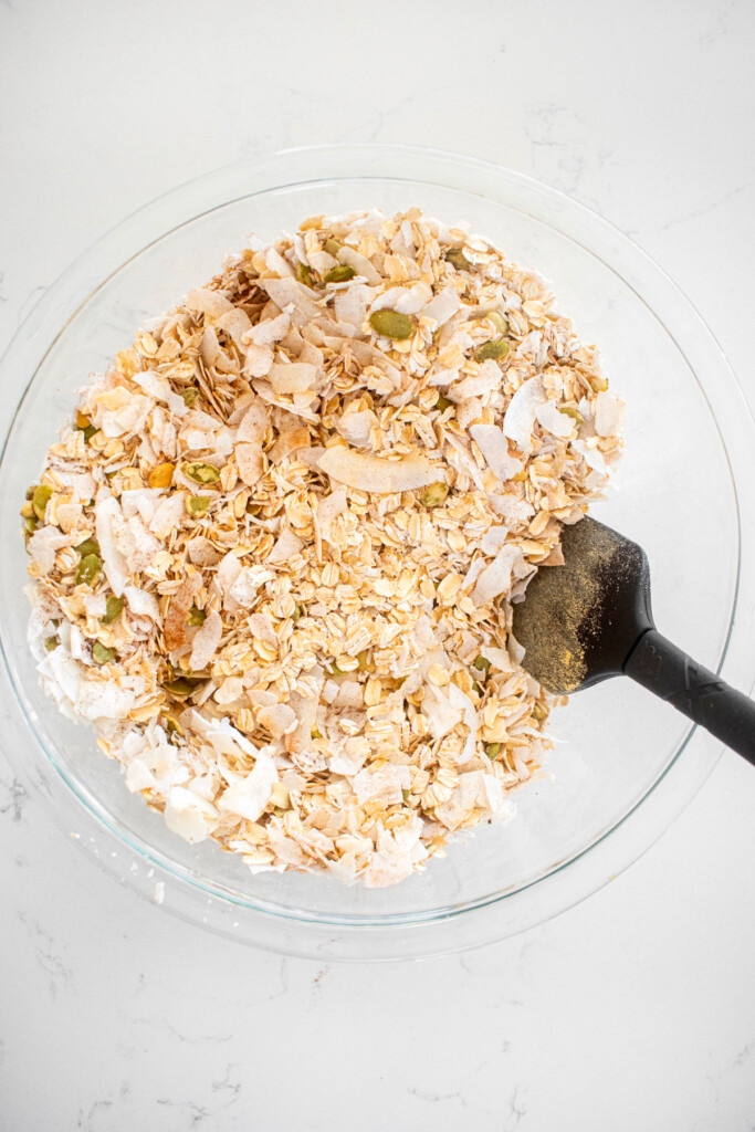 ingredients for healthy gingerbread granola mixed together in a glass mixing bowl on a white marble counter.