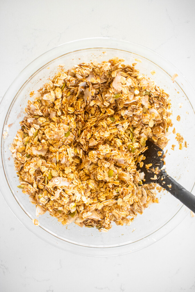 ingredients for healthy gingerbread granola being mixed together in a glass mixing bowl on a white marble counter.