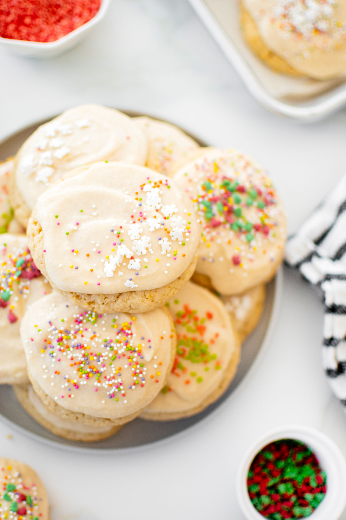 holiday sugar cookies with icing on a plate on a marble counter with sprinkles in bowls.