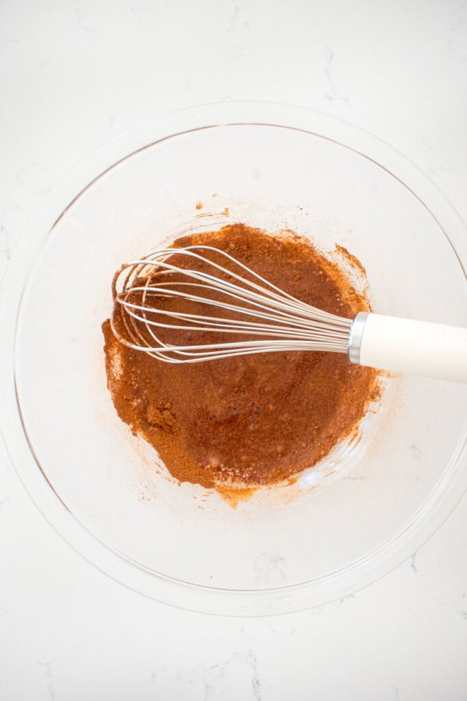 aquafaba in a glass mixing bowl mixed with cinnamon and salt with a whisk on a marble counter.