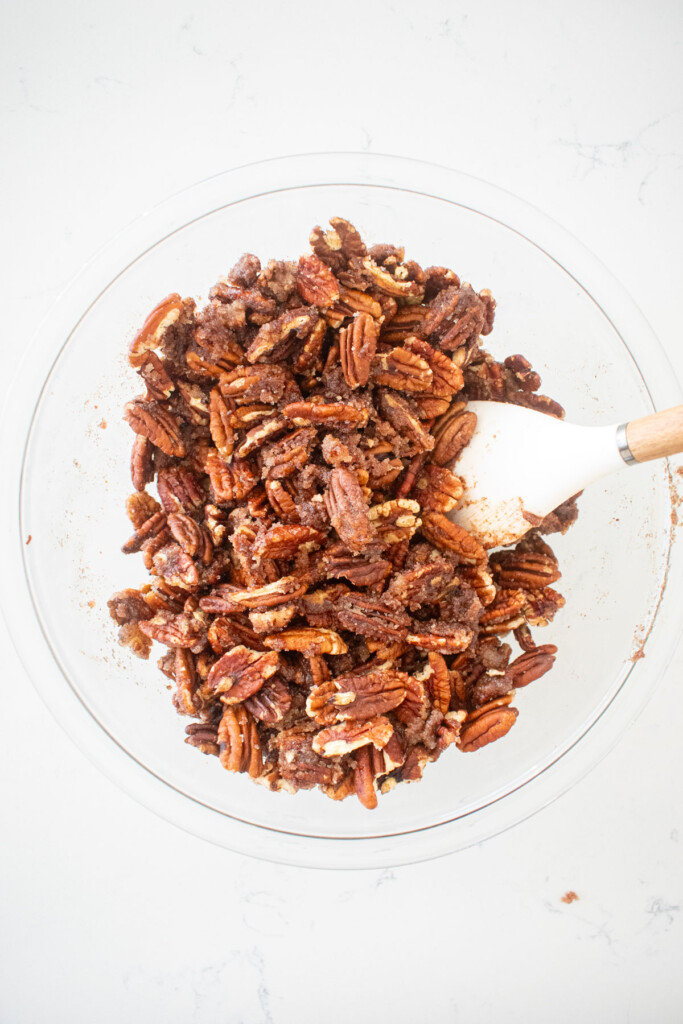 candied pecans mixed together with a spatula in a glass mixing bowl on a marble counter.