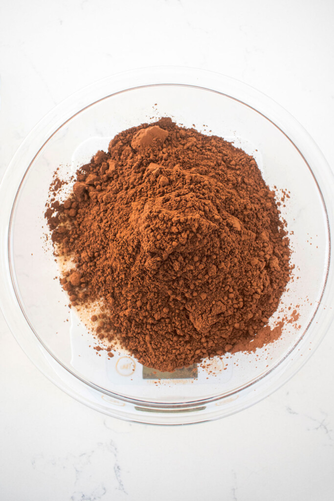 cane sugar and dutch cocoa powder in a glass mixing bowl on top of a scale on a white marble counter.