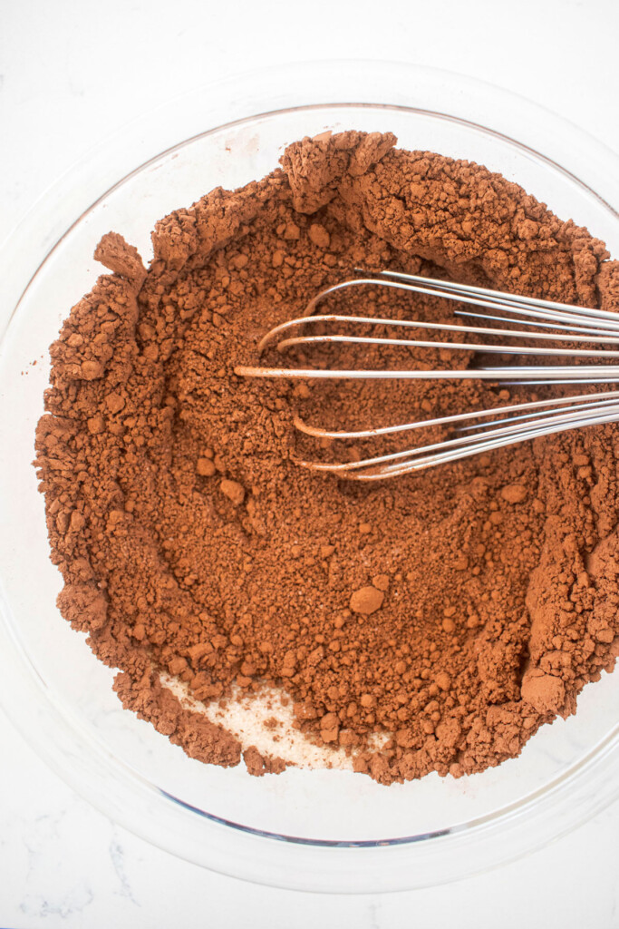 homemade hot cocoa mix being mixed together in a glass mixing bowl with a whisk on a white marble counter.