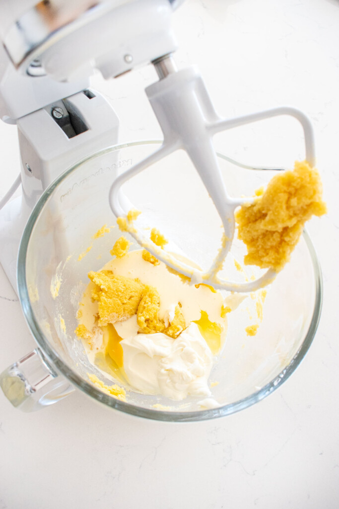 creamed butter and sugar with eggs, yogurt, and cream in a glass mixing bowl on a white marble counter.