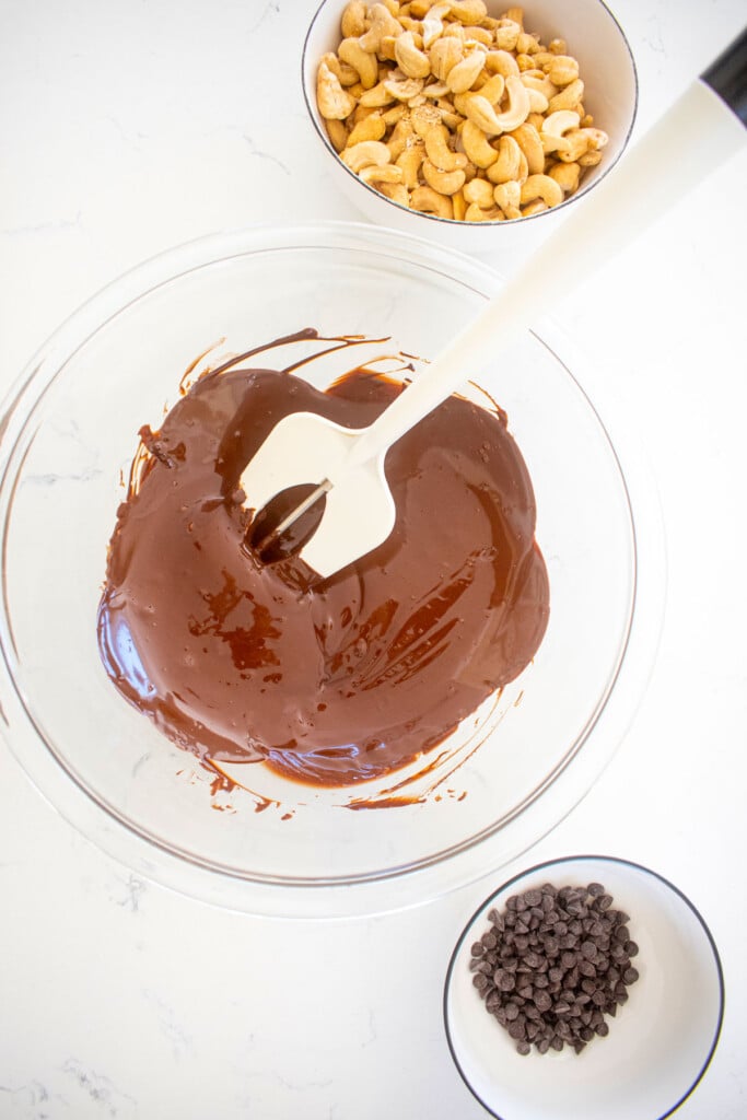melted chocolate in a glass mixing bowl with a bowl of chocolate chips and cashews on the side on a marble counter.