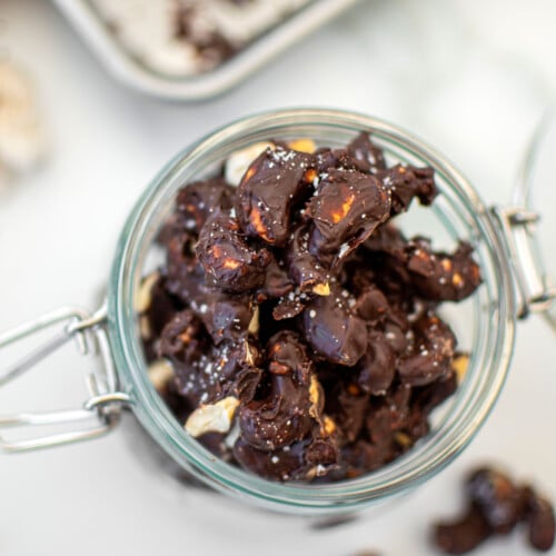 sea salt dark chocolate cashew clusters in a glass jar on a white marble counter.