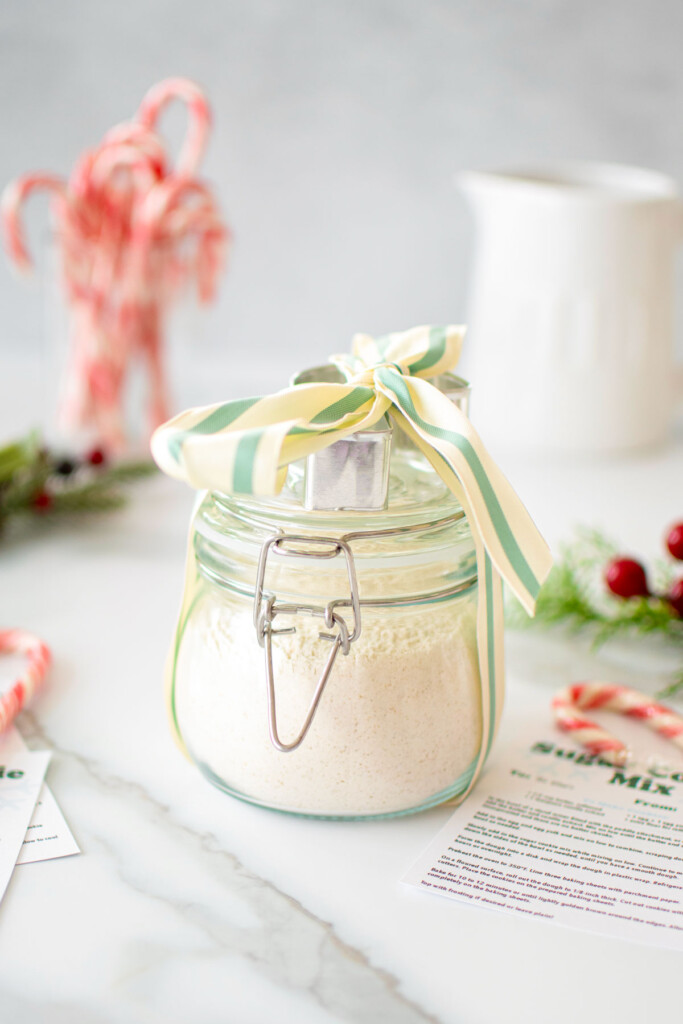 sugar cookie mix for gifts in a glass jar with a cookie cutter on top and tied with a ribbon on a marble counter.