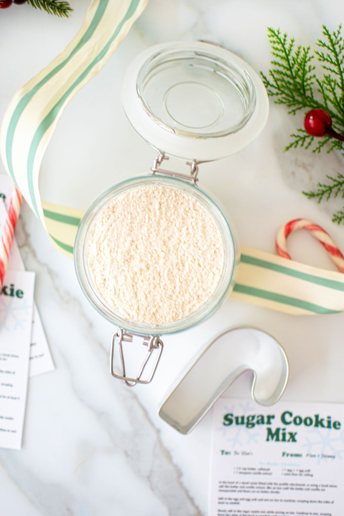 sugar cookie mix for gifts in a glass jar on a marble counter with holiday ribbon candy canes and cookie cutter.