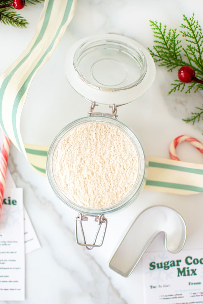 sugar cookie mix for gifts in a glass jar on a marble counter with holiday ribbon candy canes and cookie cutter.
