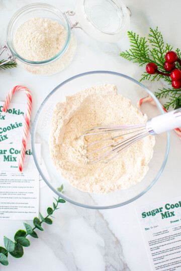 sugar cookie mix for gifts being mixed in a glass mixing bowl with a whisk on a marble counter with ribbons and candy canes.