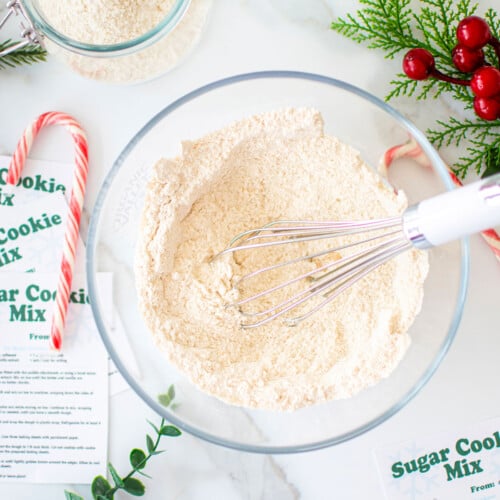 sugar cookie mix for gifts being mixed in a glass mixing bowl with a whisk on a marble counter with ribbons and candy canes.