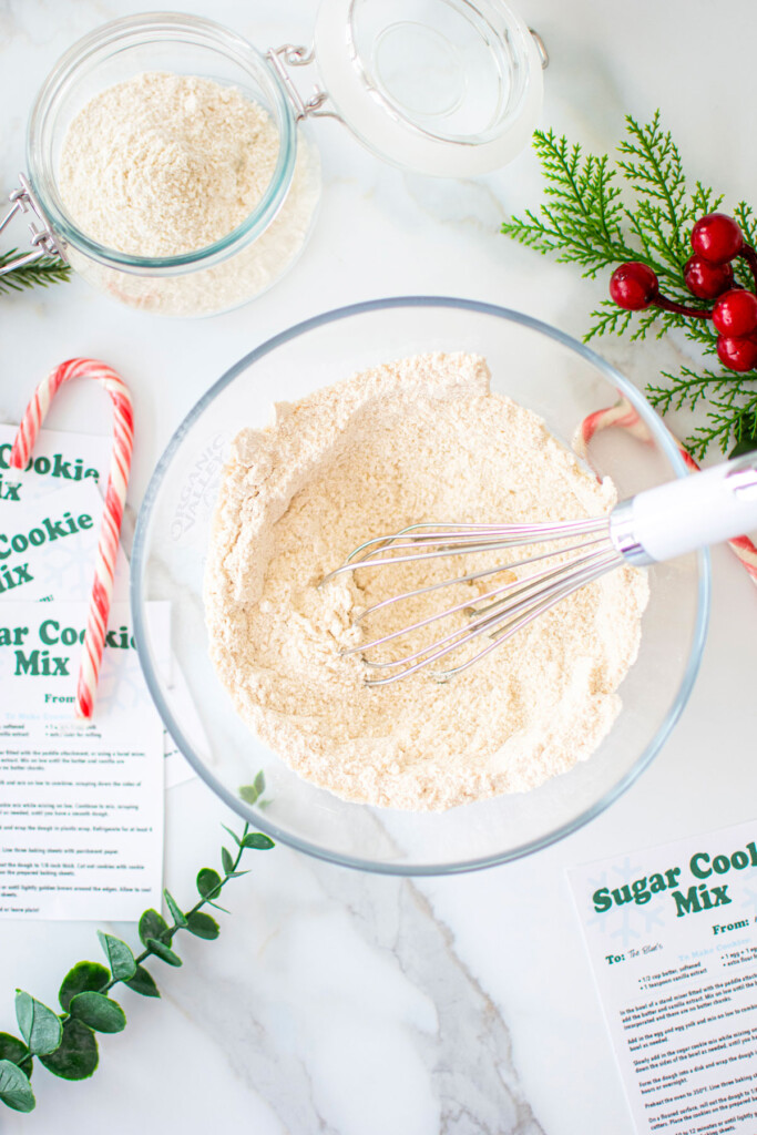 sugar cookie mix for gifts being mixed in a glass mixing bowl with a whisk on a marble counter with ribbons and candy canes.