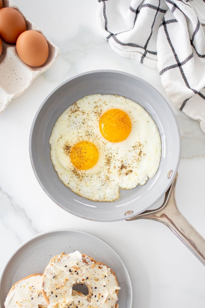 caraway pan with 2 fried eggs in it on the counter with a bagel on a plate.